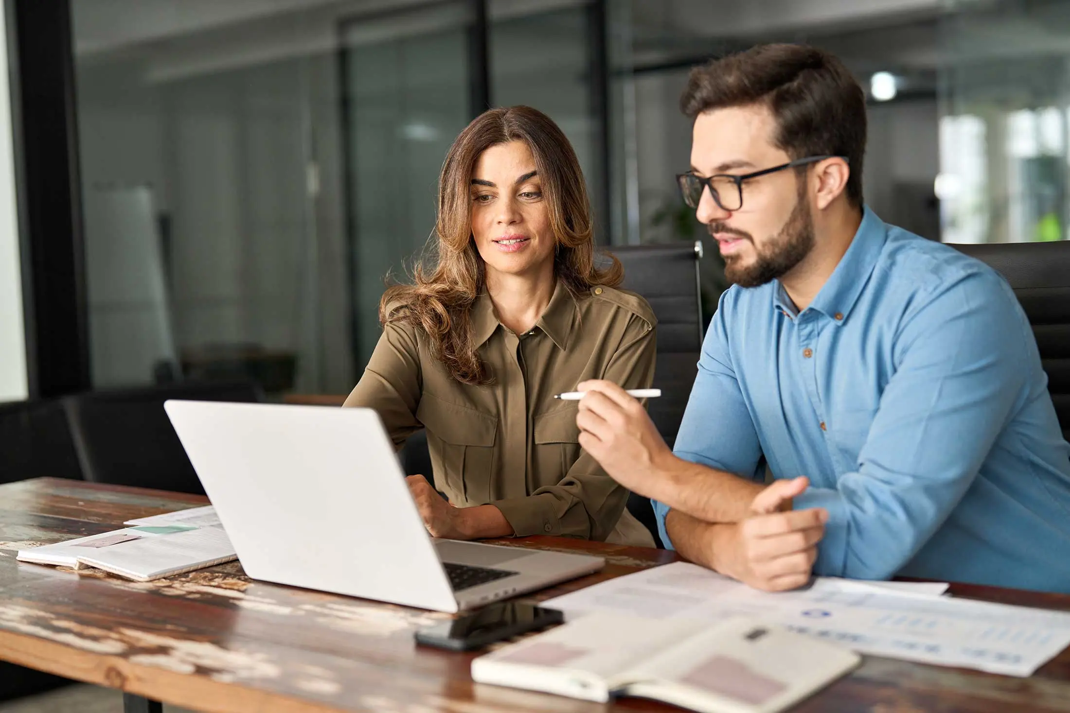 Een man en een vrouw zitten aan een tafel en werken samen aan een laptop. Ze zijn in gesprek, wisselen ideeën uit en bekijken de resultaten van SustainQ.